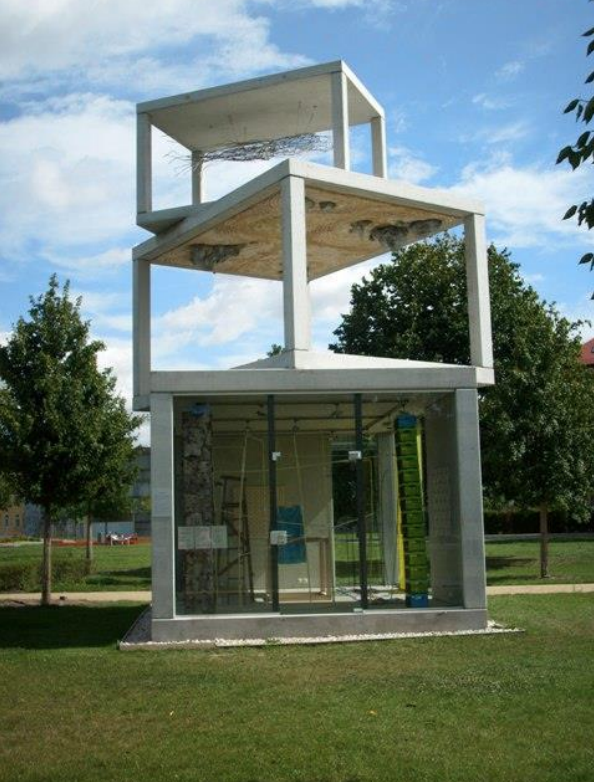 Skulptur aus drei gestapelten Betonrahmen, darunter verglaster Raum, auf einer Rasenfläche mit Bäumen und blauem Himmel.